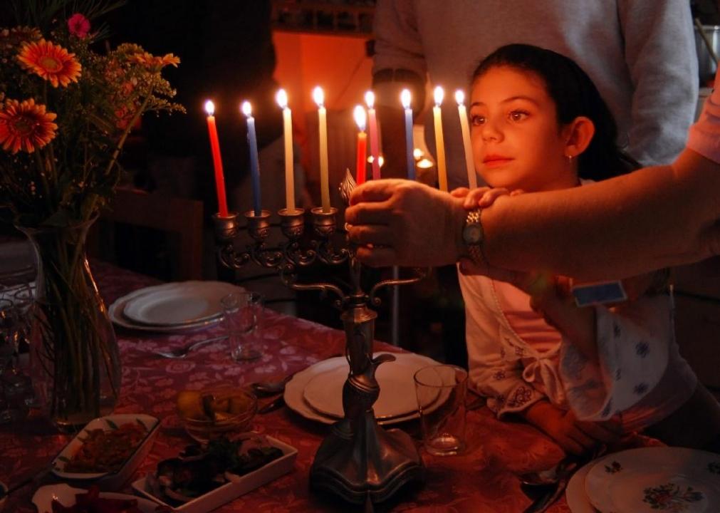 A young child looks at a menorah being lit at a dining table.