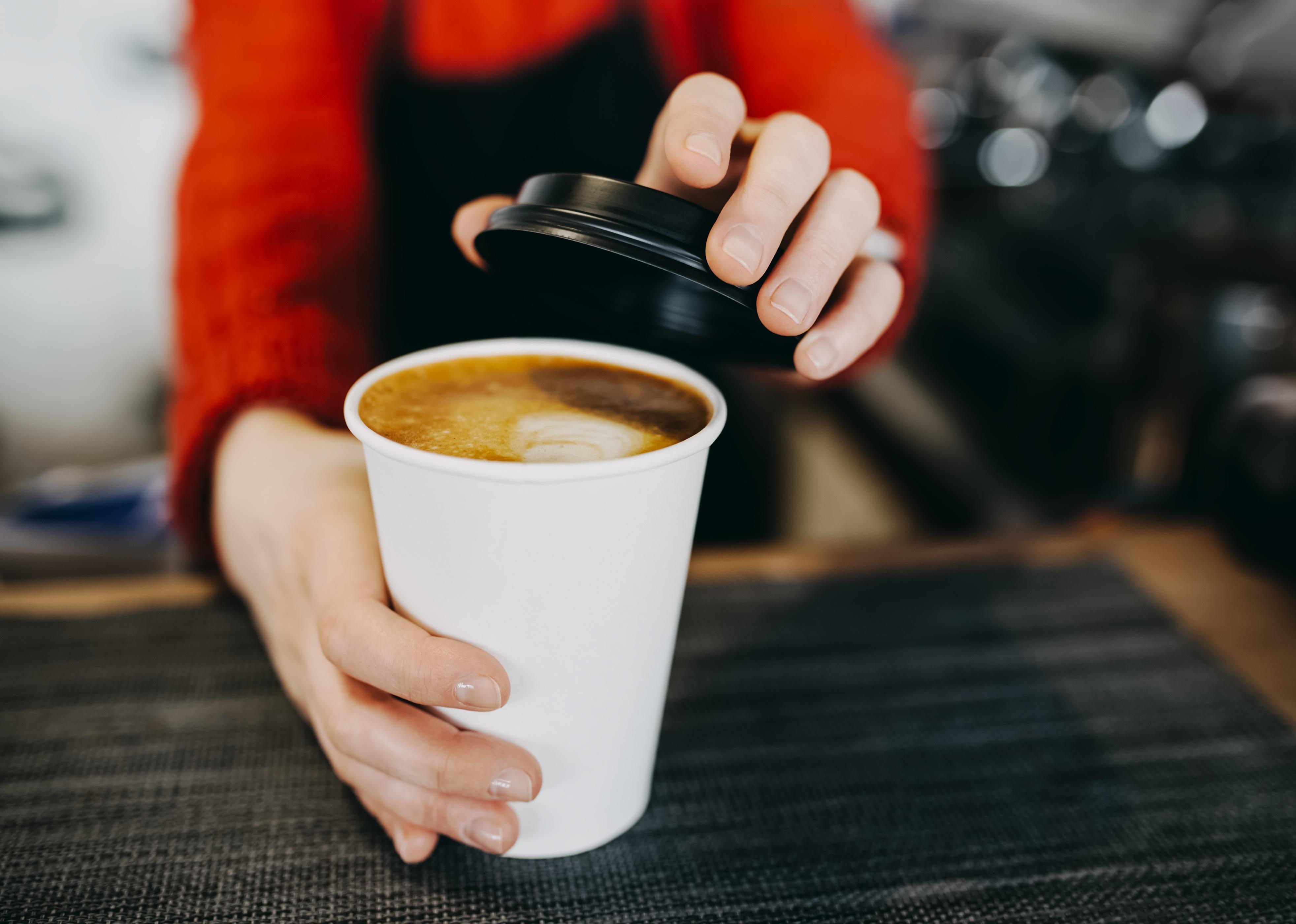 Barista in apron is holding a hot cappuccino in white to go paper cup. 