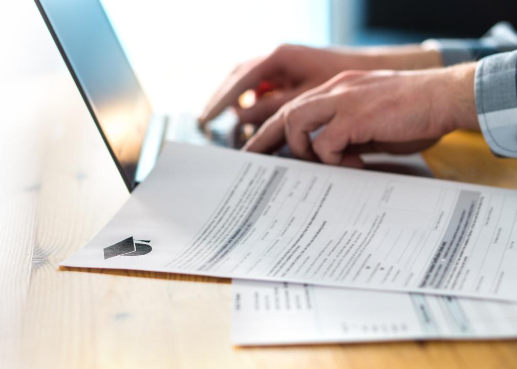 A person typing on a laptop with paper college forms also on the desk. 