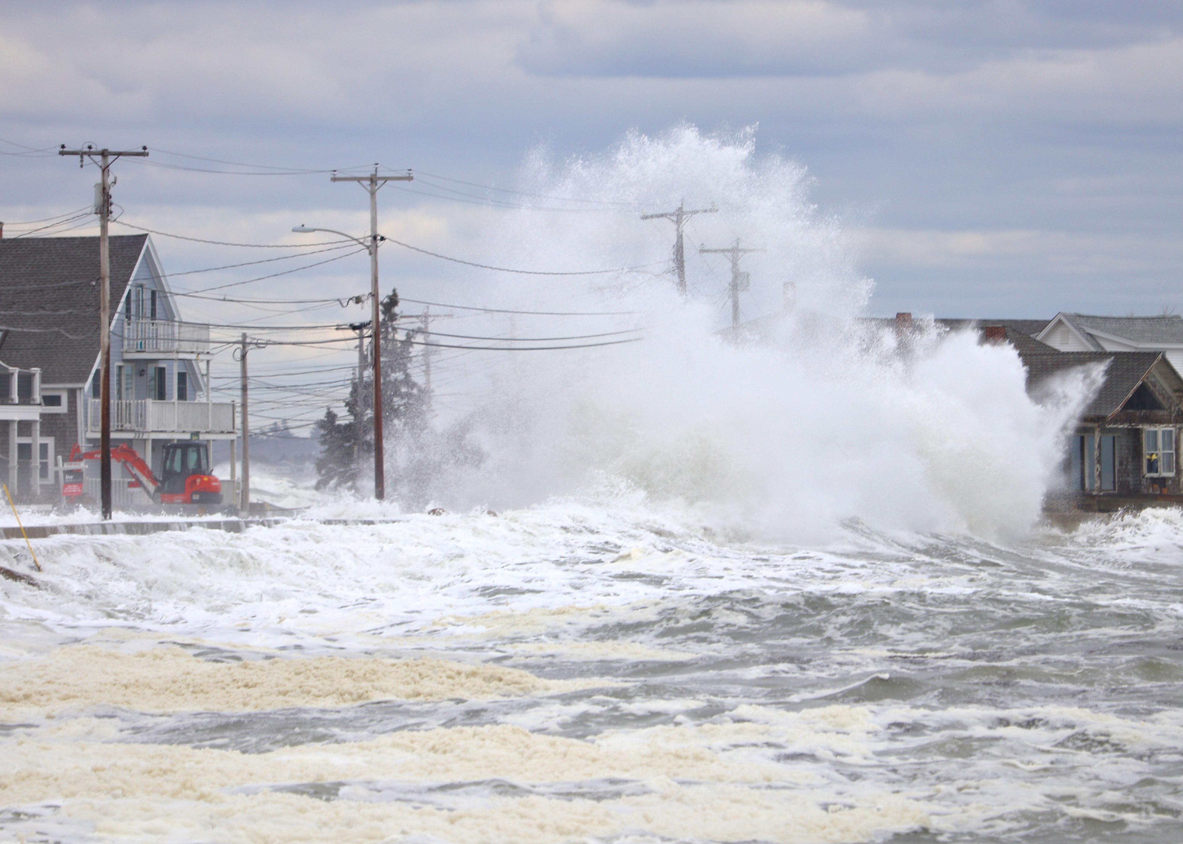 A storm wave crashes over two telephone poles and the roof of a beach front house.