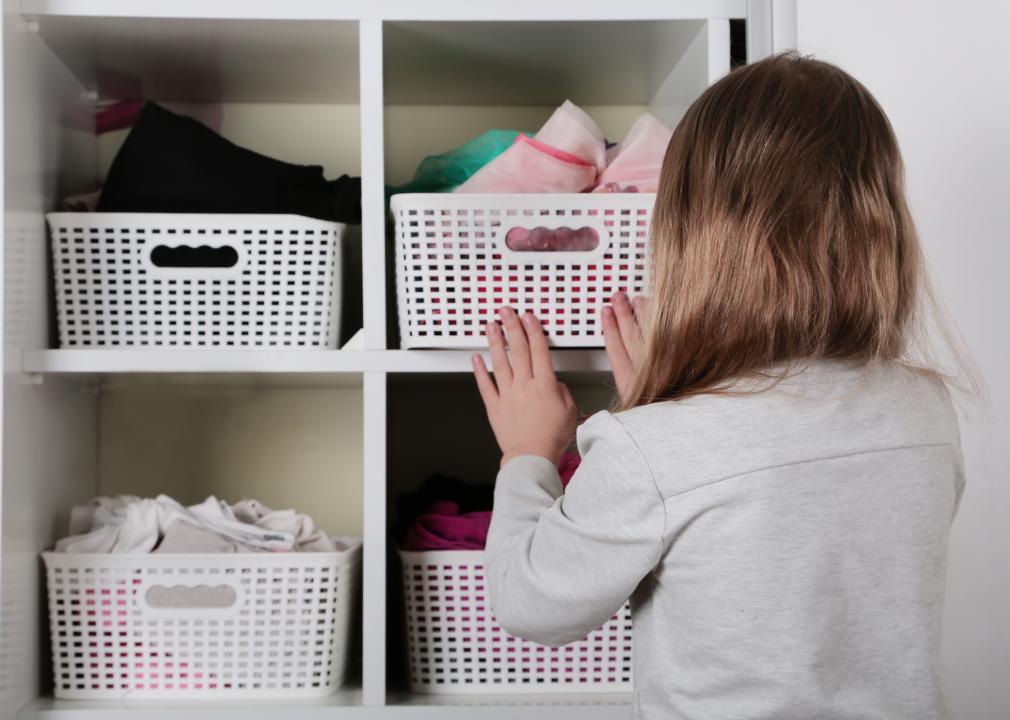 A child puts away folded clothes inside of baskets. The baskets are placed on shelves inside of a closet.