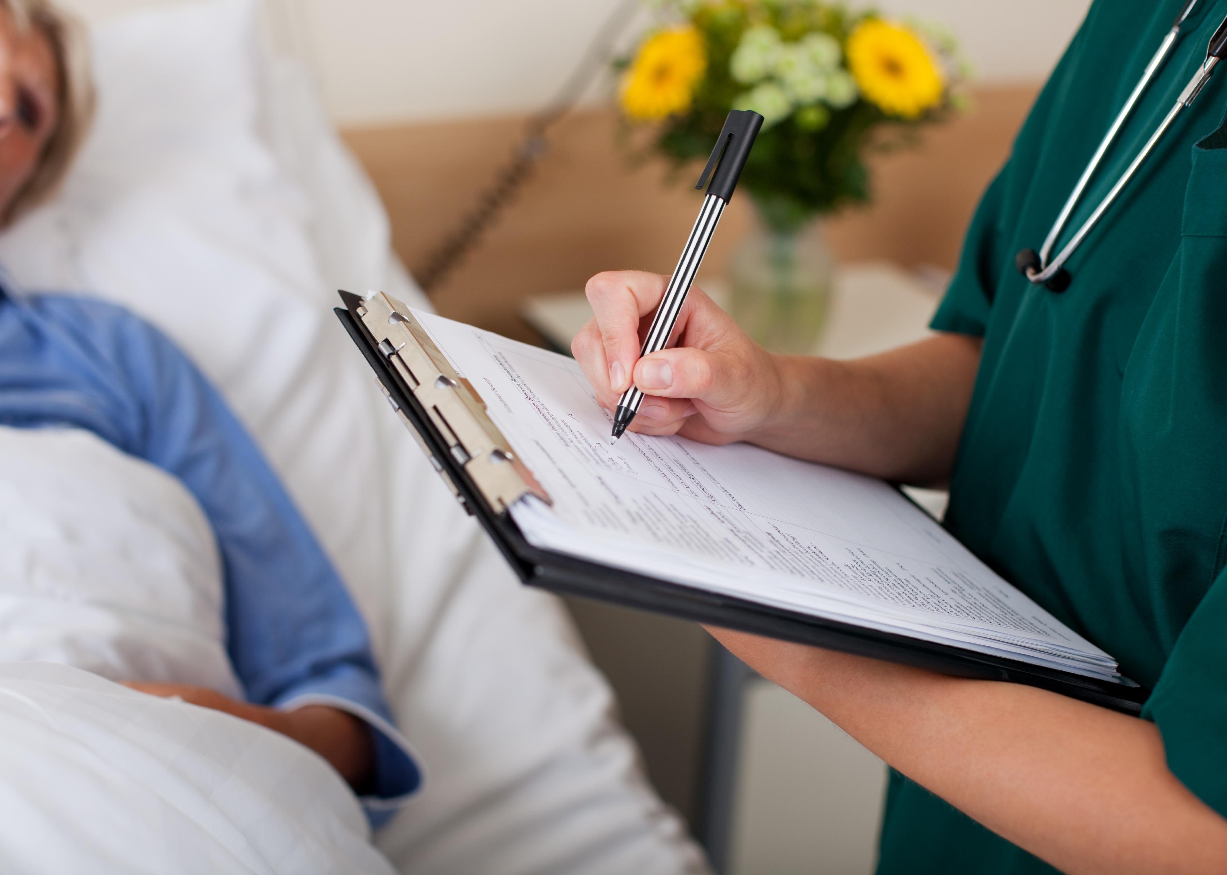 Nurse writing on clipboard with patient in background in a hospital bed.