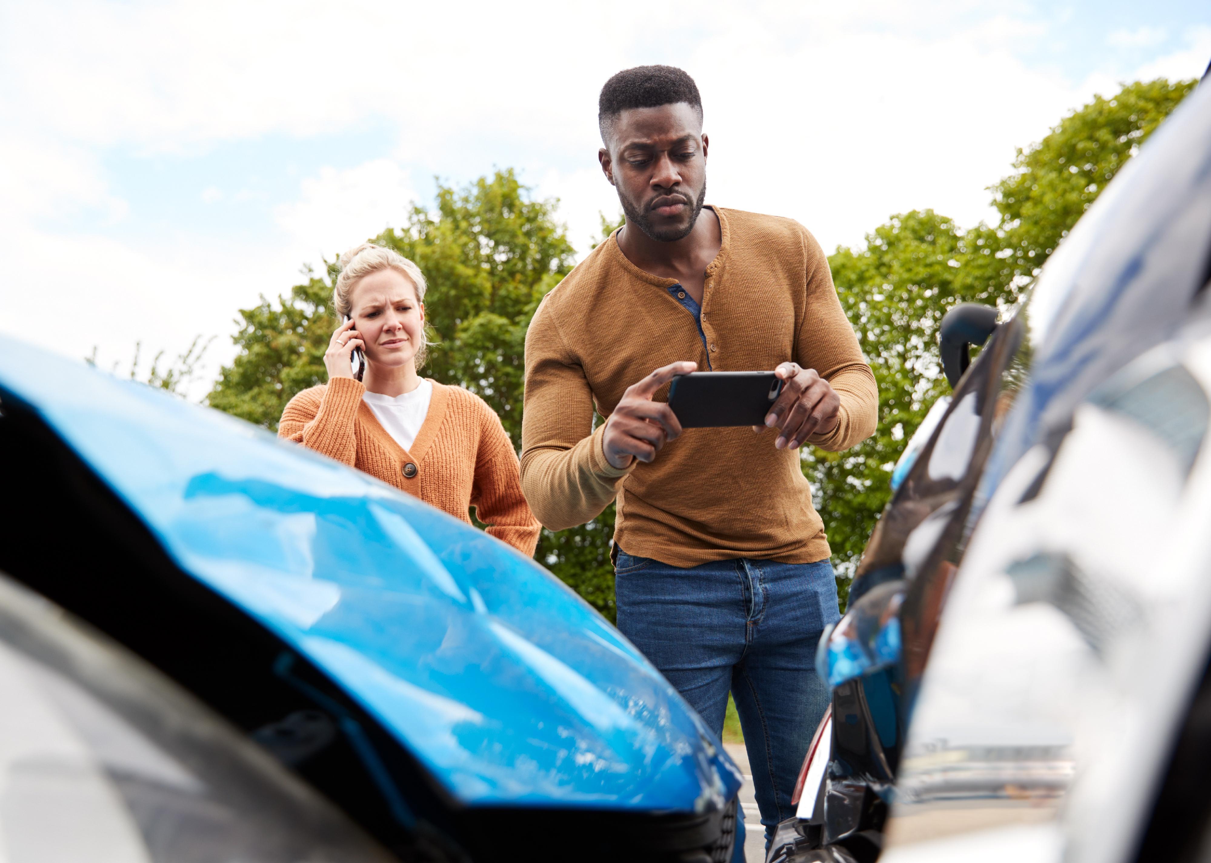 A man involved in car accident taking a picture of damage for an insurance claim while a woman is on the phone behind him..