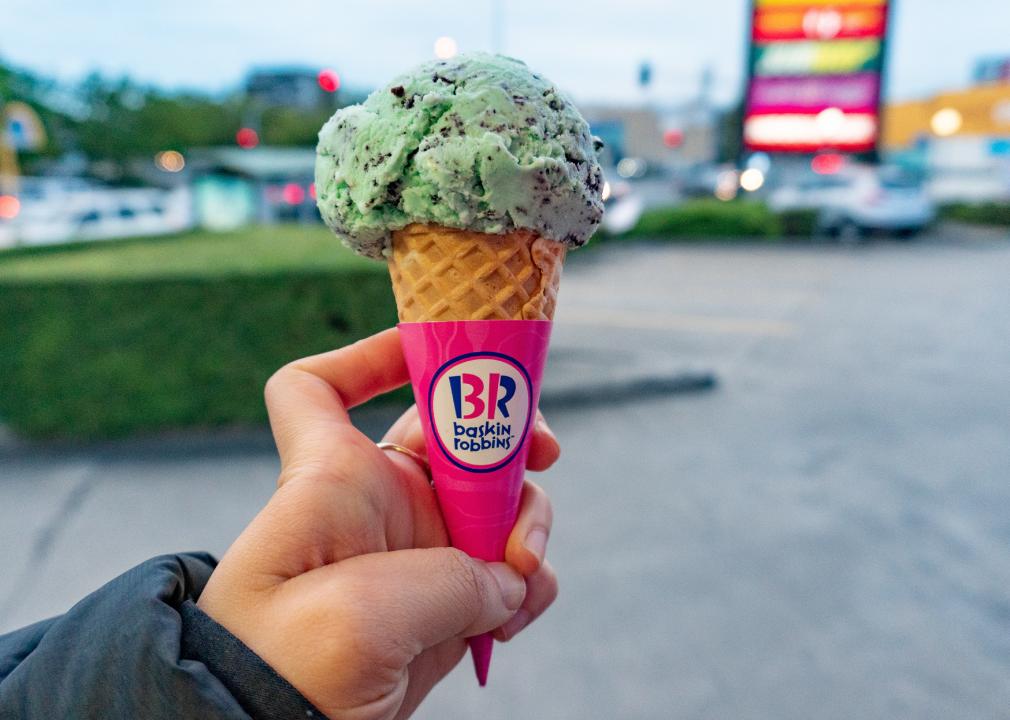 A close up of a person's hand holding a Baskin Robbins cone with a scoop of mint chocolate chip ice cream. 