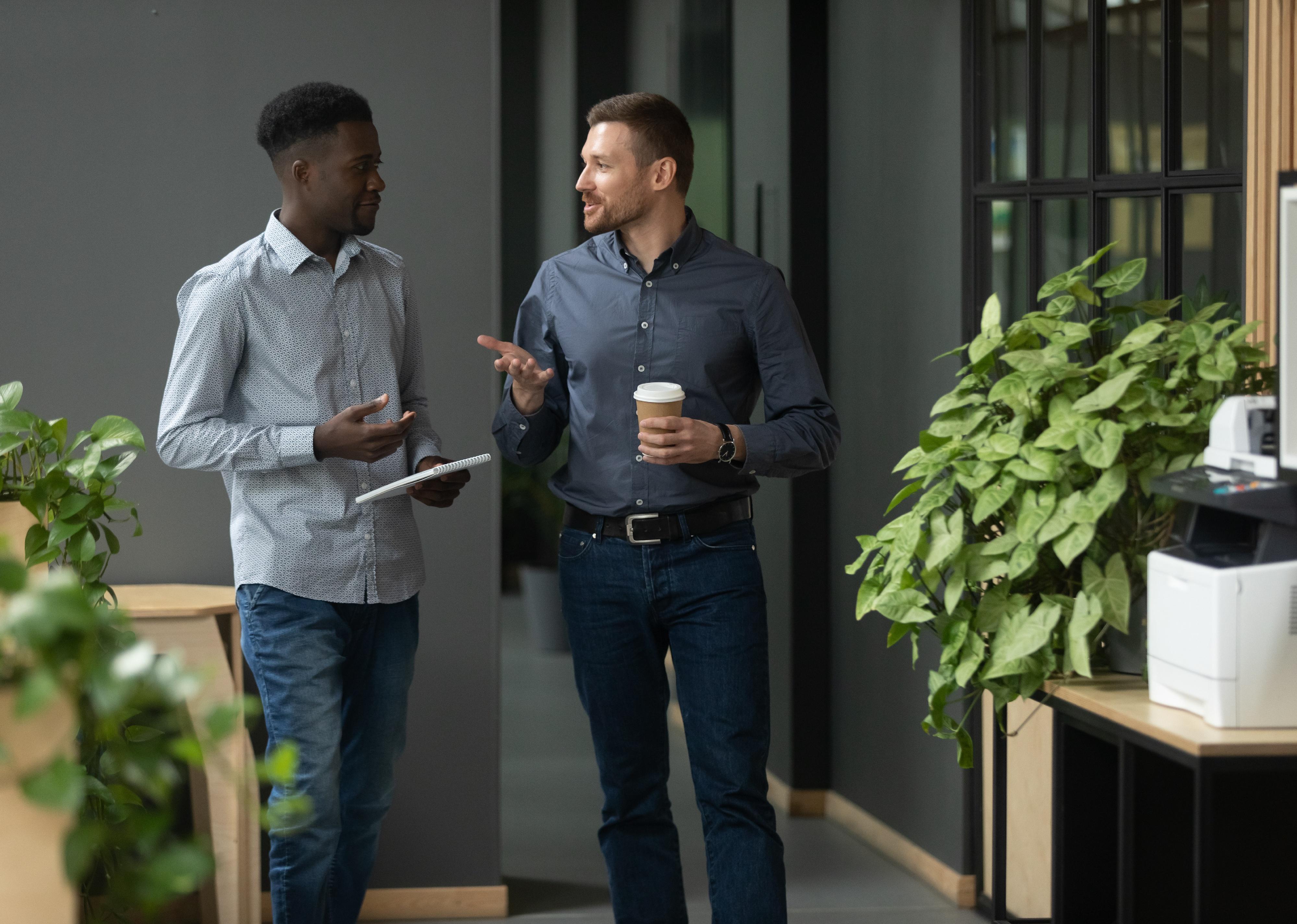 Two male colleagues talking and walking in modern office hallway.