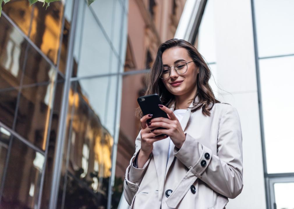 Smiling woman wearing sunglasses walks down a city street and uses her phone.