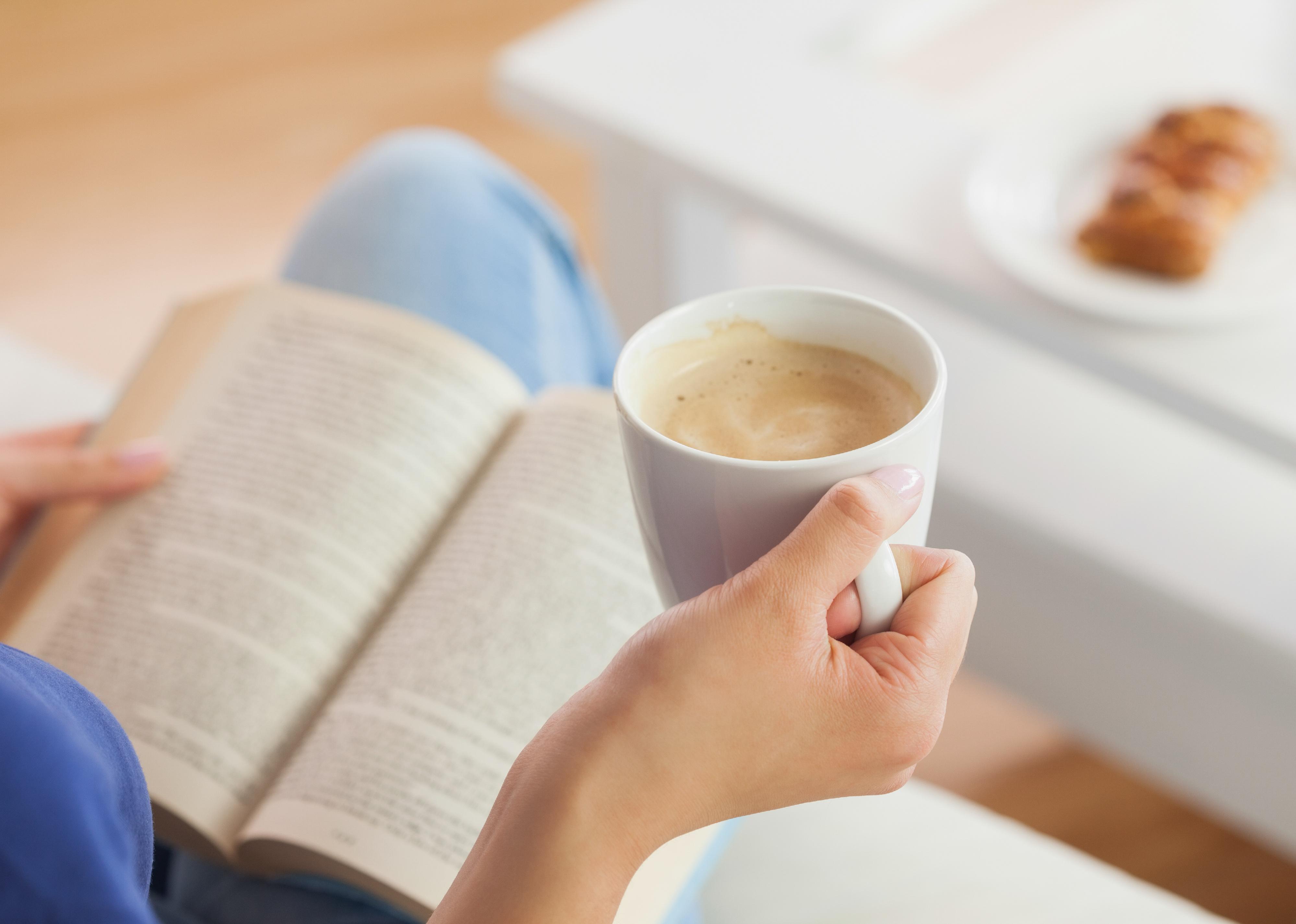 Woman sitting on the sofa, reading a book, and holding a coffee mug at home.