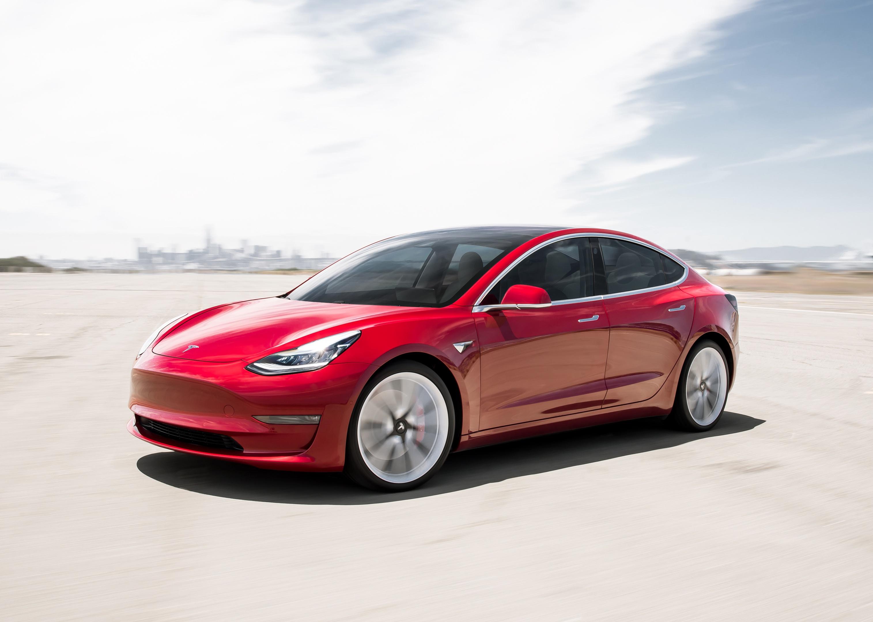 A red Tesla Model 3 in front of a distant city.