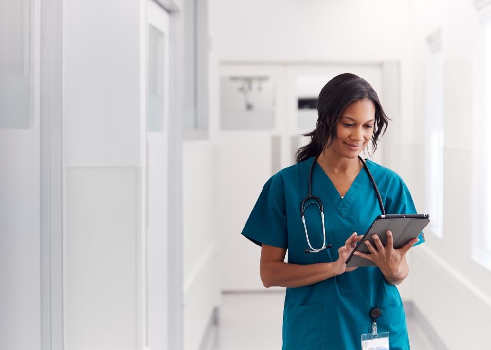 A nurse wearing scrubs walks in hospital corridor as she looks at a tablet.