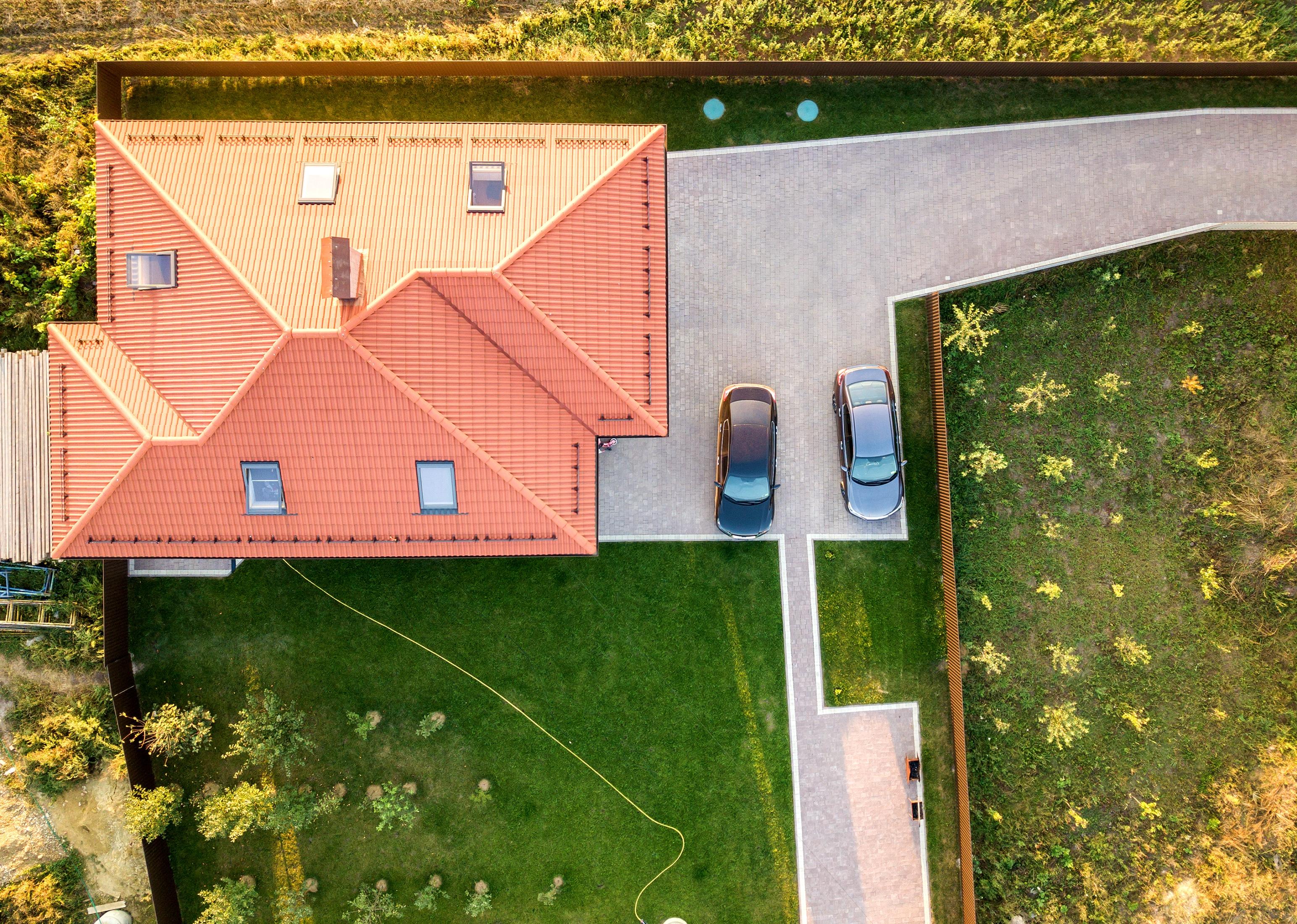 Top-down aerial view of a house with red roof and yard with two parked new cars.