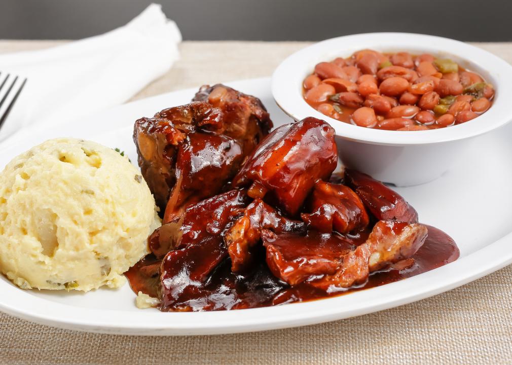 A plate of barbecue rib tips with sauce with a bowl of beans and a scoop of mashed potatoes.