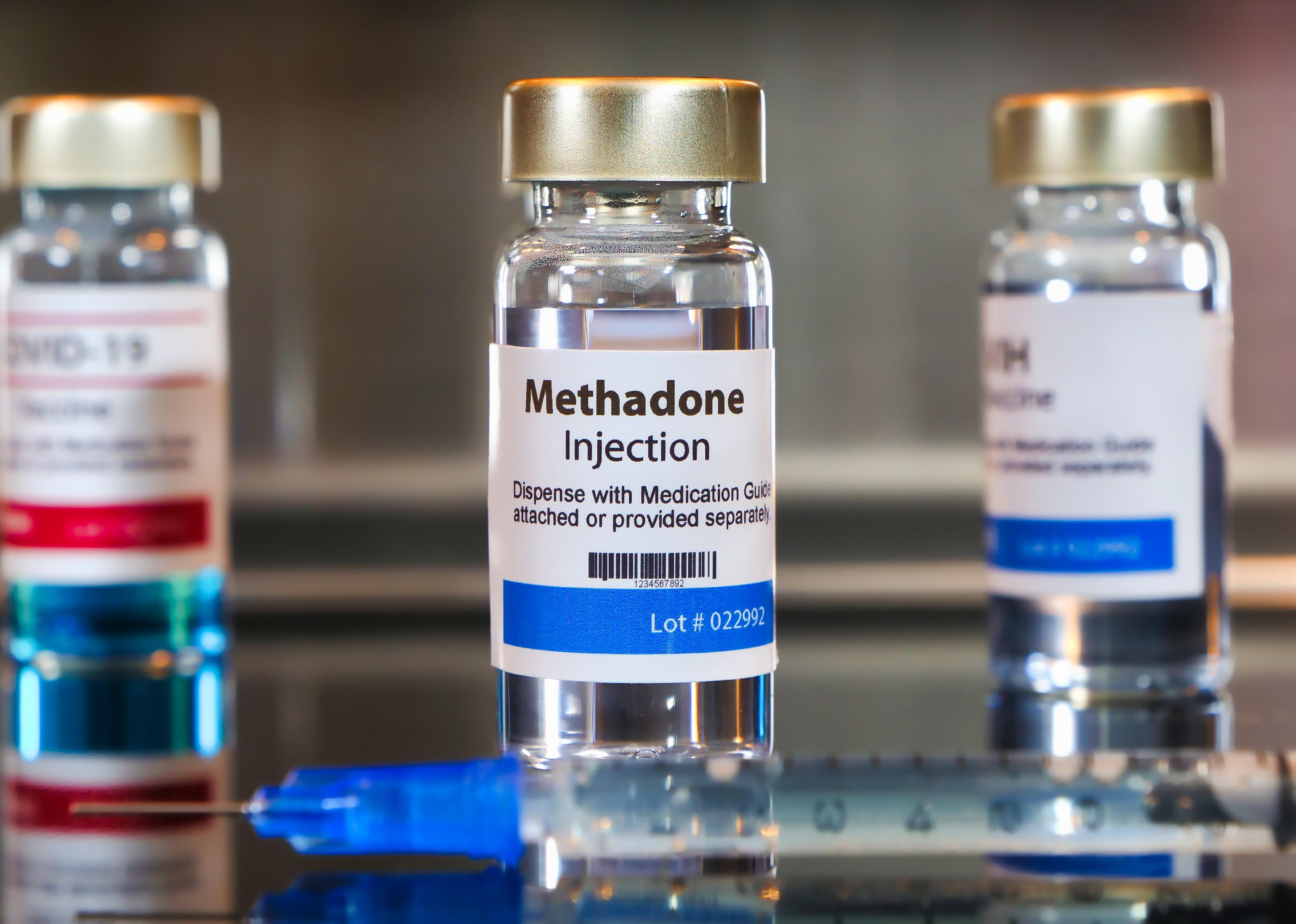 Vial of methadone injection with a syringe on black table and stainless steel background.