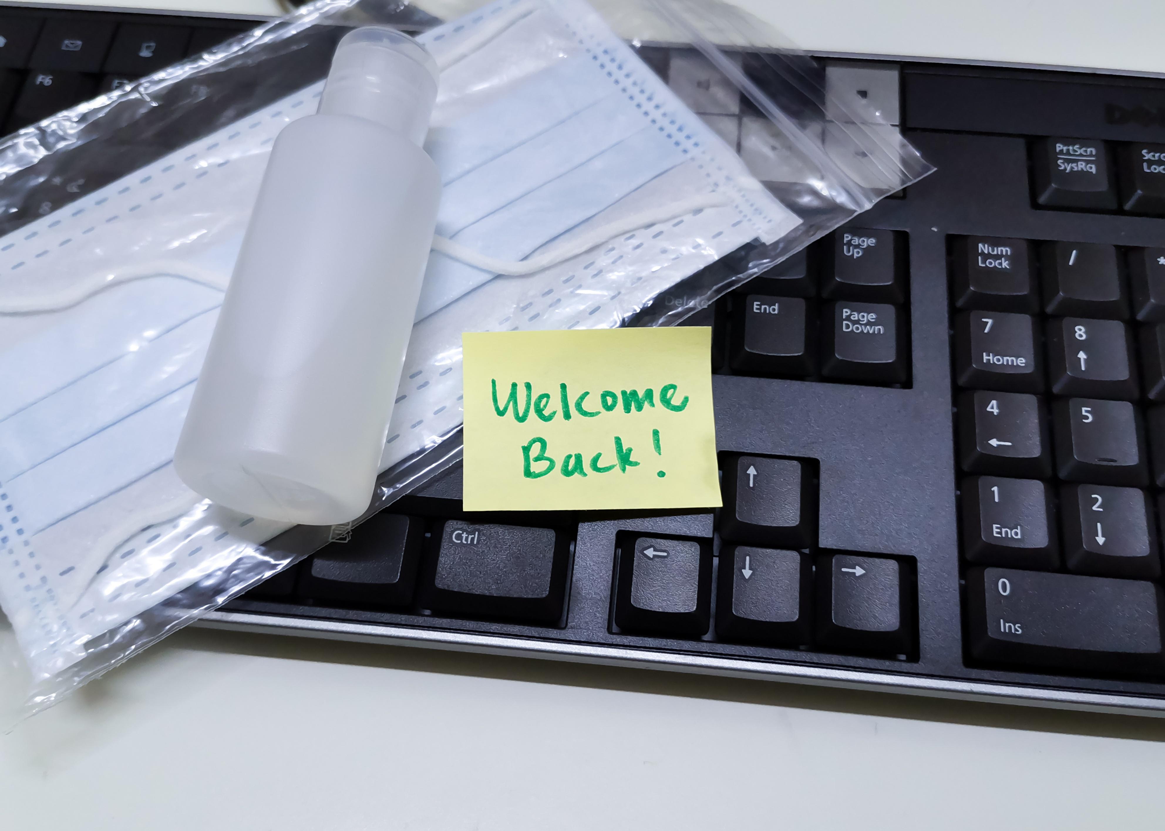 Welcome back note with hand sanitizer and mask on work keyboard.