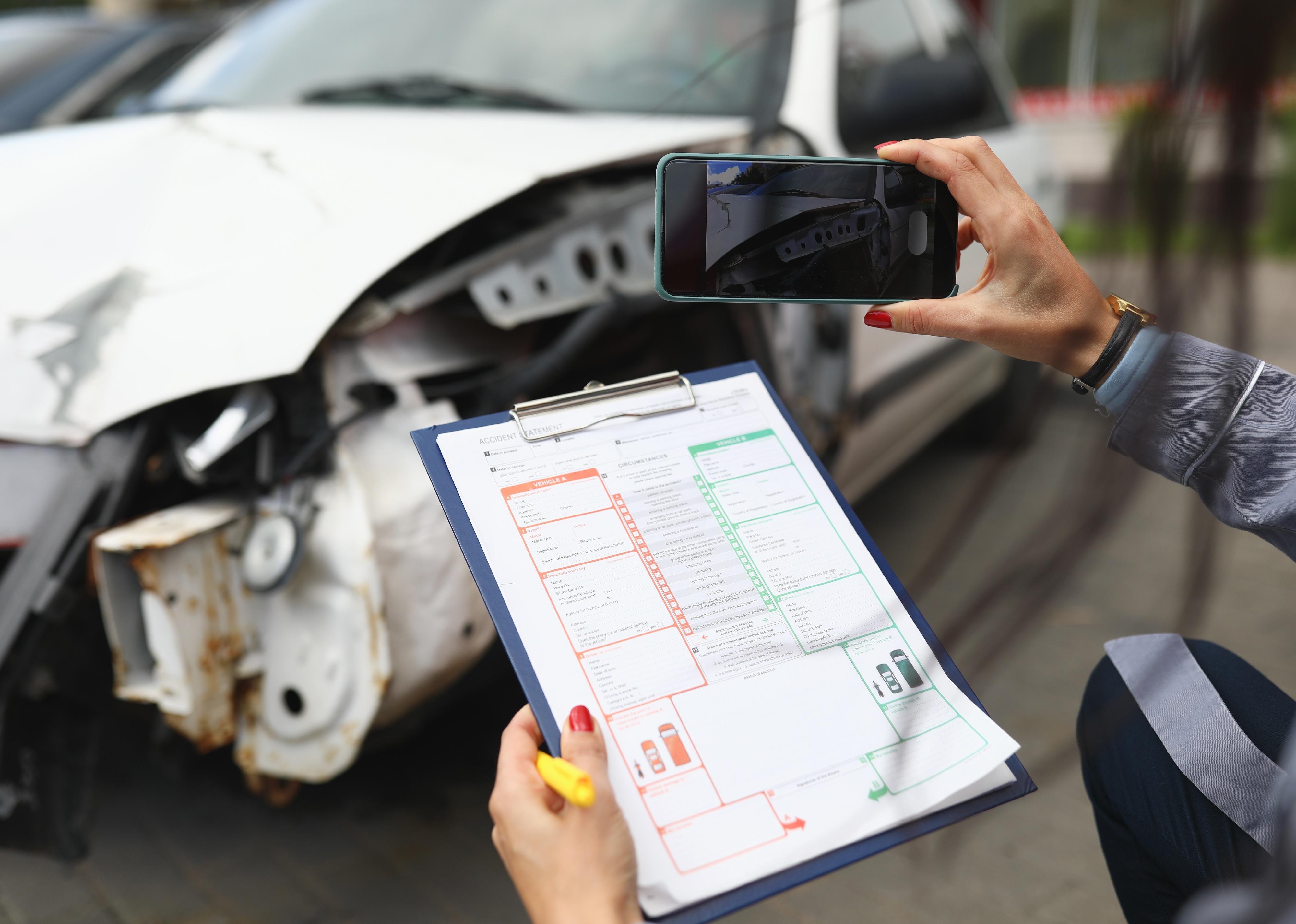 A woman photographs a broken car on a smartphone and holds insurance documents in her hands