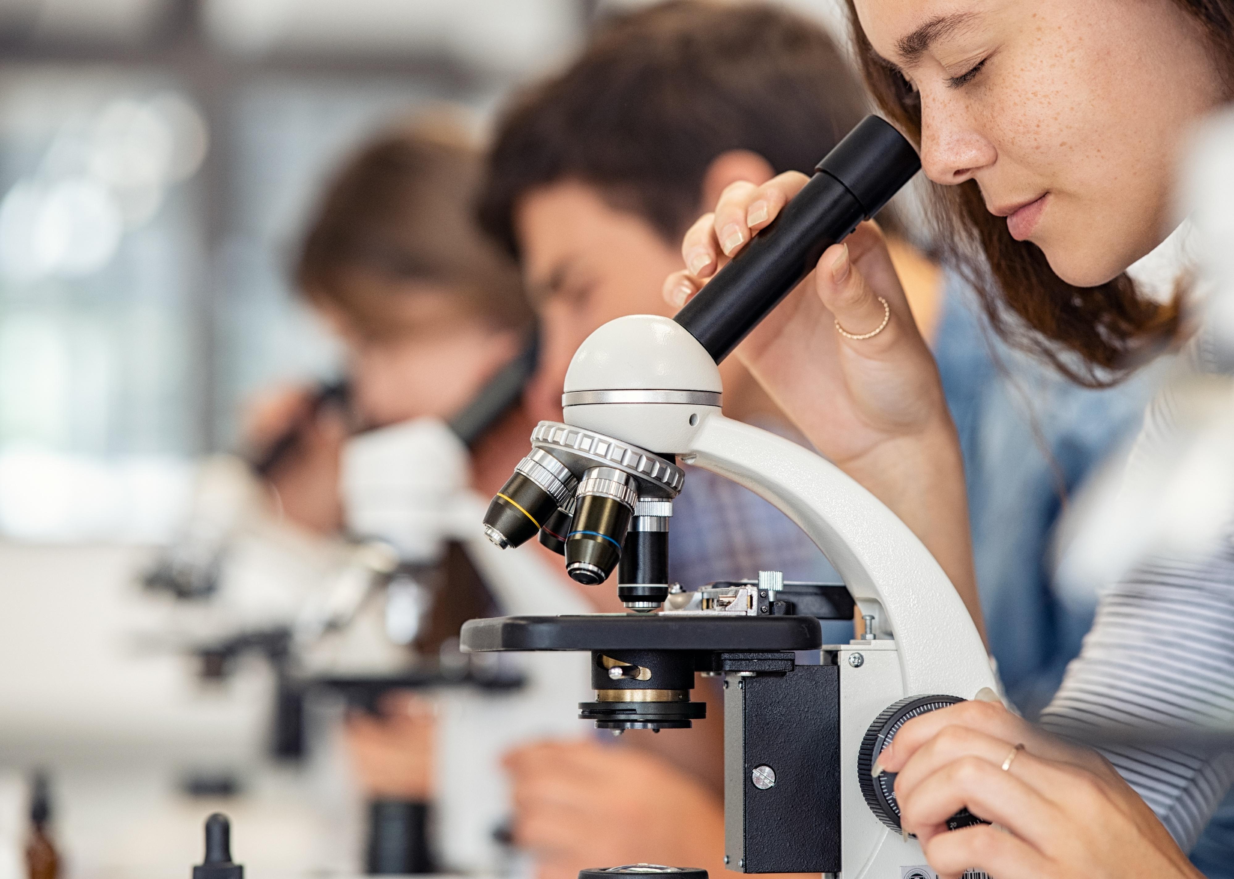 Close up of young woman looking through microscope with other students