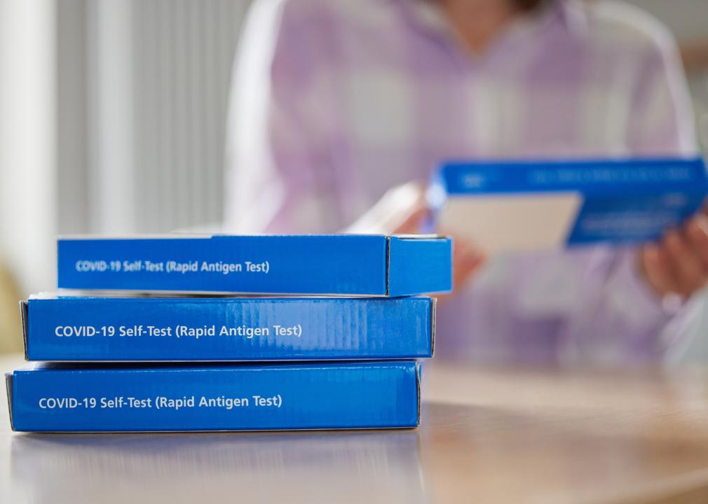 Three boxes of COVID-19 Rapid Antigen Self-Testing Kits with a woman in the background reading the instructions on another box.