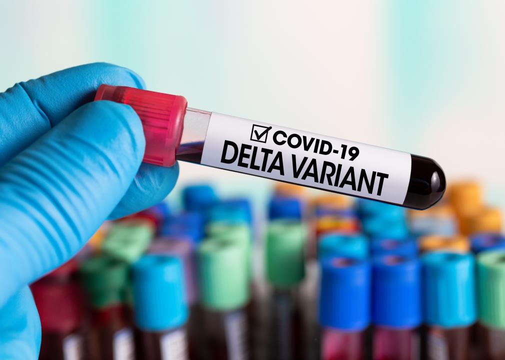 A hand wearing a blue glove holding a tube of blood labeled COVID-19 Delta Variant.