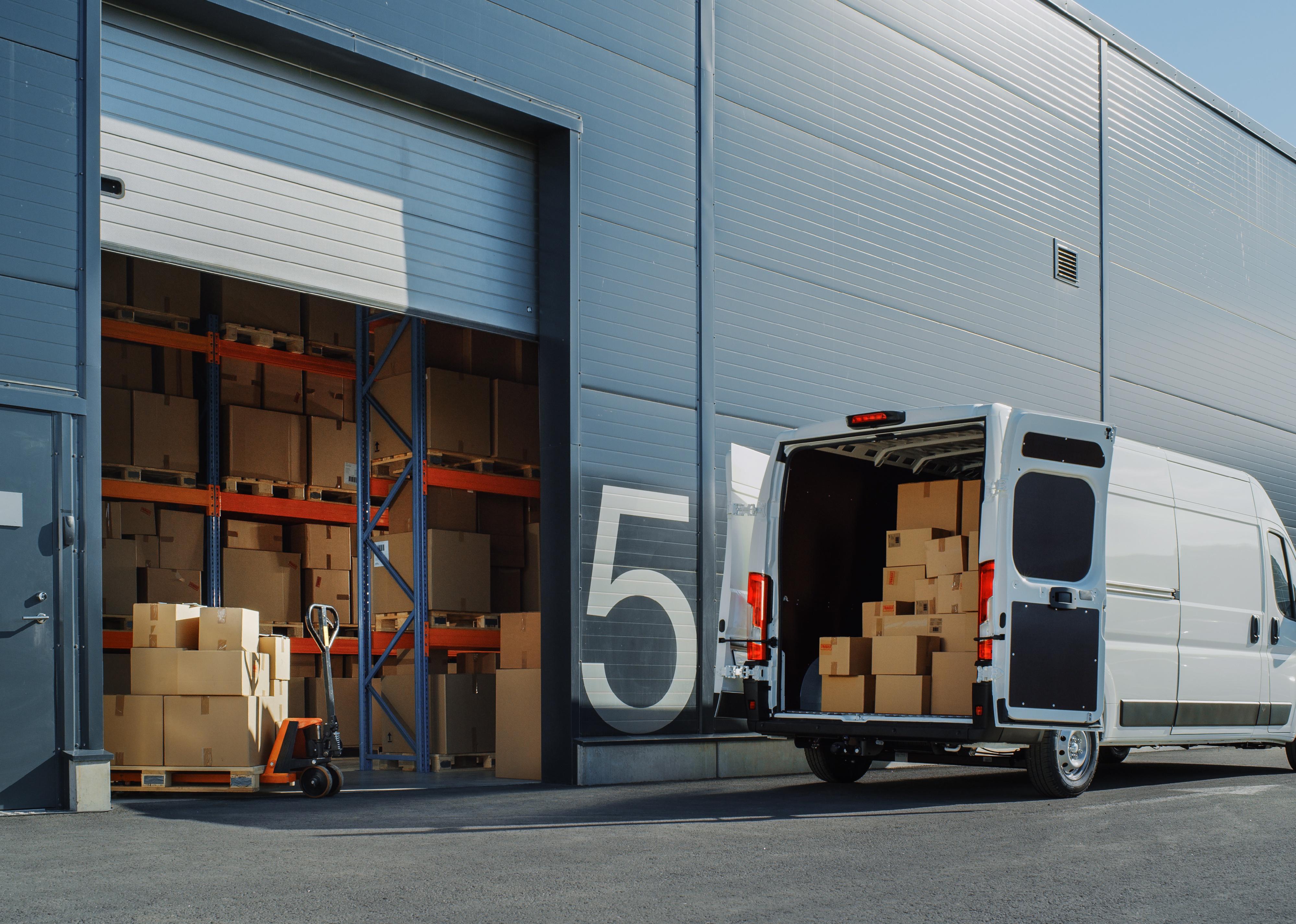 Delivery van loaded with cardboard boxes outside of logistics warehouse with open door.