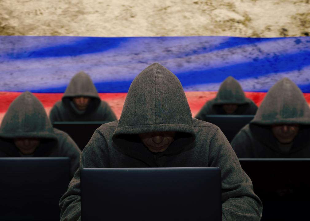Many hackers are sitting at laptops and computers against the background of the Russian flag