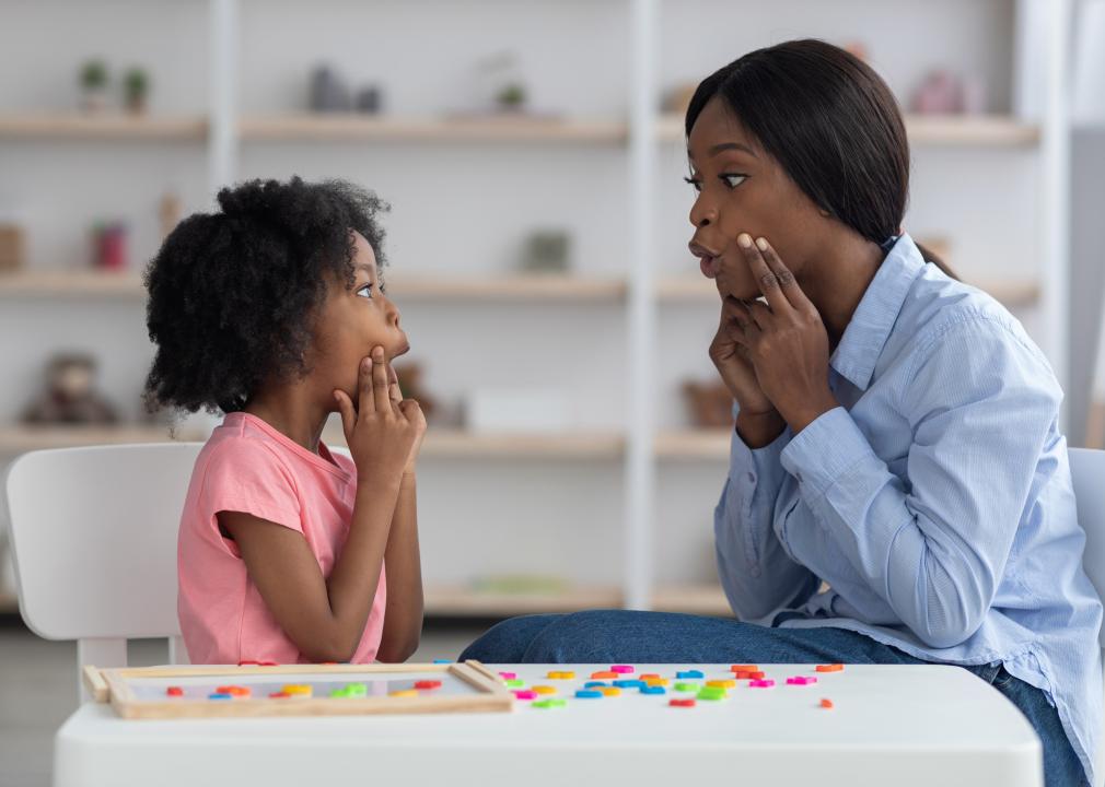 A speech-language pathologist having a lesson with a young child. They are both holding their hands to their cheeks.