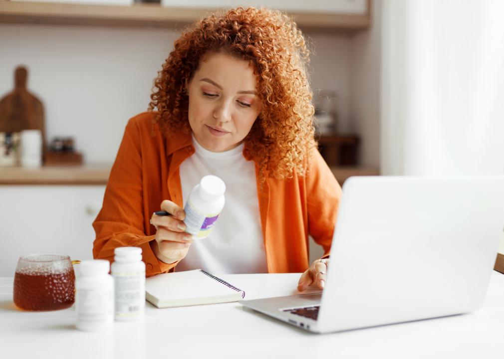 Woman on laptop sitting at kitchen table with vitamin bottle.