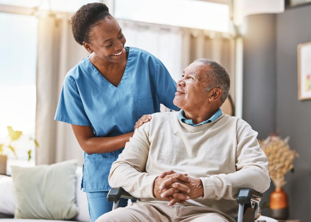 A smiling nurse stands with her hands on the shoulders of a smiling patient in a wheelchair.