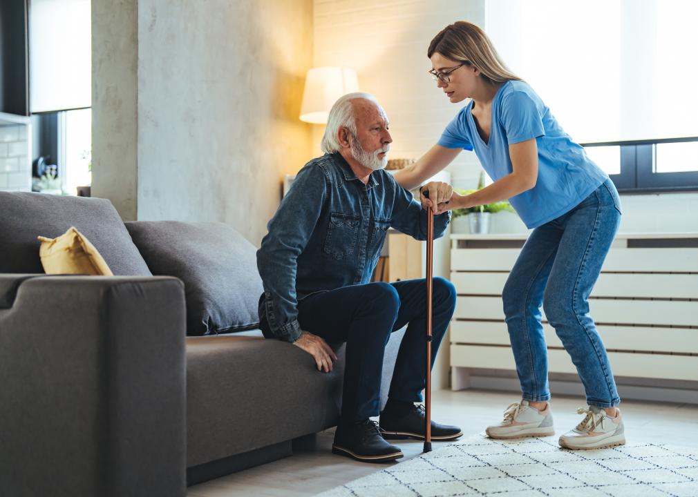 A home aid helping a senior man with a cane stand up from a couch.