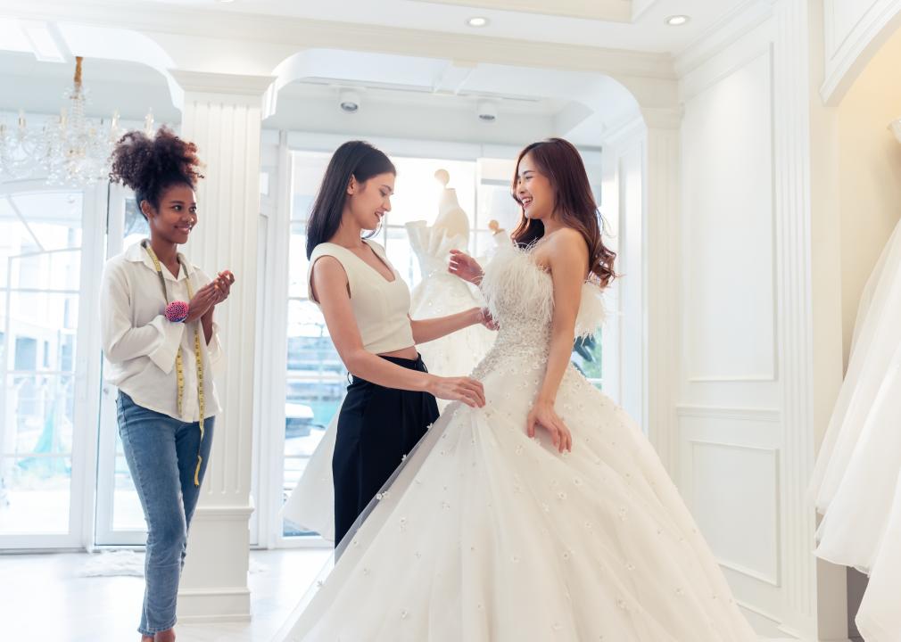 A woman trying on a wedding gown in a shop; one woman is adjusting the dress and another is standing next to them with a tape measurer hanging around her neck.