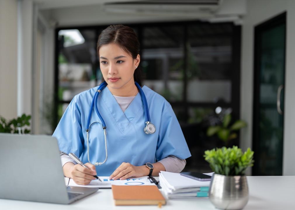 A health care worker in scrubs stands in front of a laptop as she writes on a piece of paper.