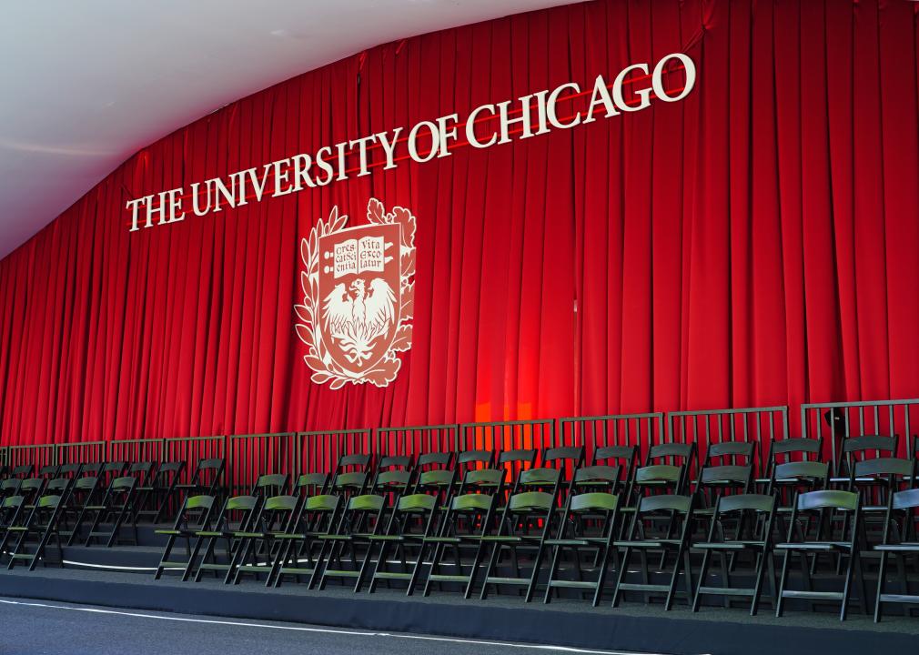 Graduation preparations, folding chairs set up in rows in front of a red curtain with the words, University of Chicago, and the university's coat of arms.