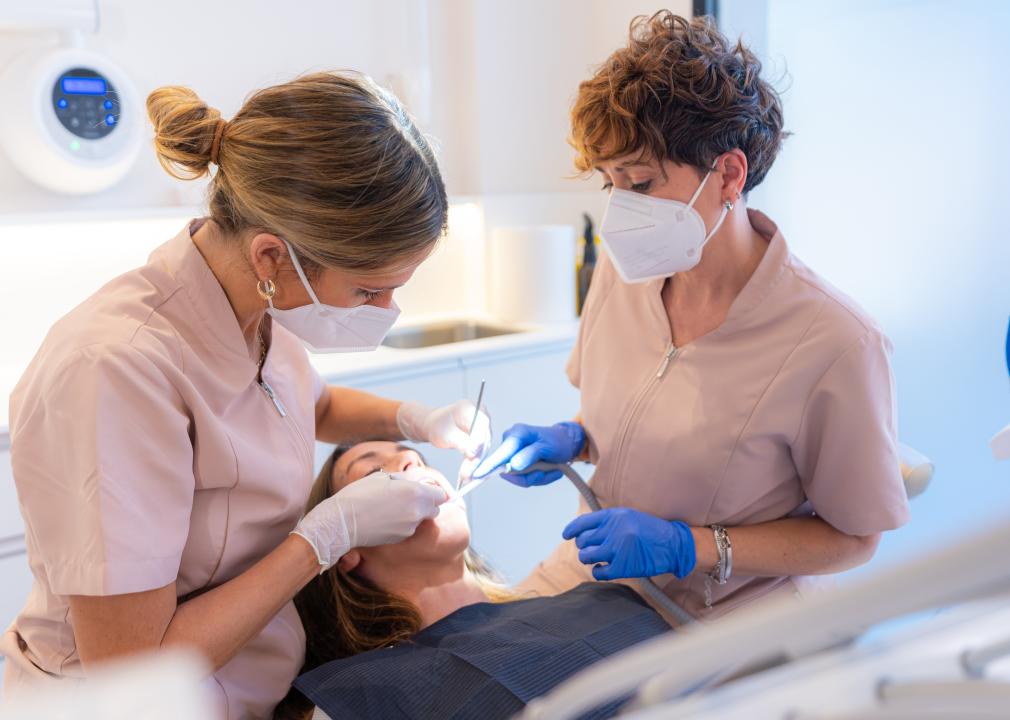 In a clinic, two dental hygienists examine the mouth of a patient sitting in a chair.