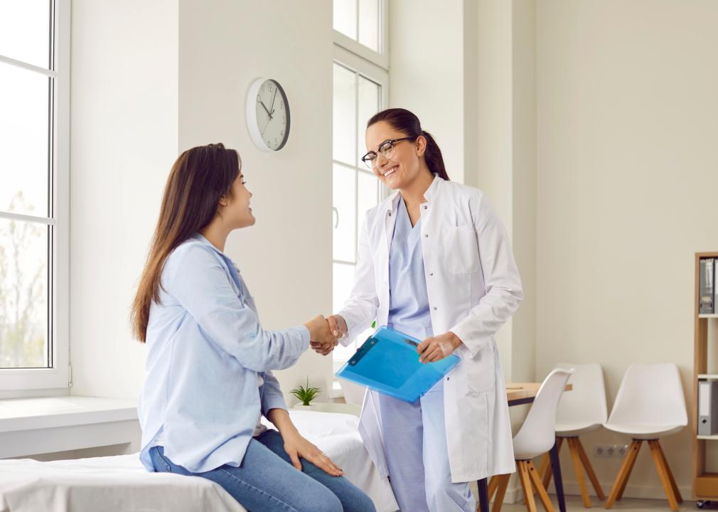 A smiling doctor shakes hands with a patient who is sitting on a bed in a medical office.