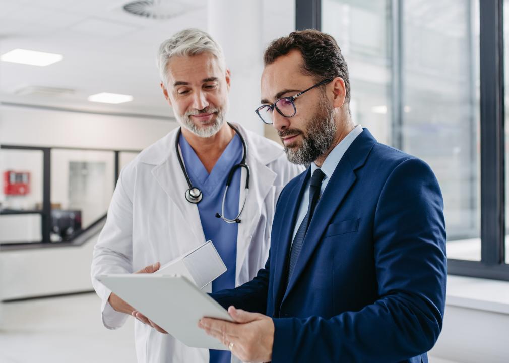 A doctor stands next to a person in a suit holding a tablet.