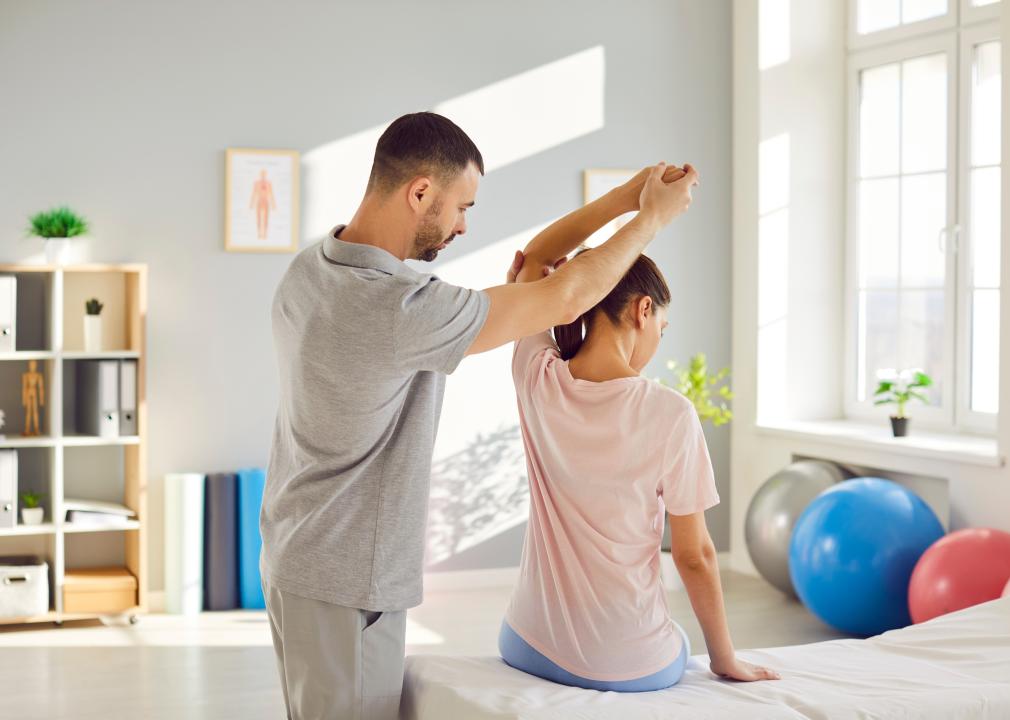 In a clinical space, a physical therapist helps a patient stretch their arm over their head. 