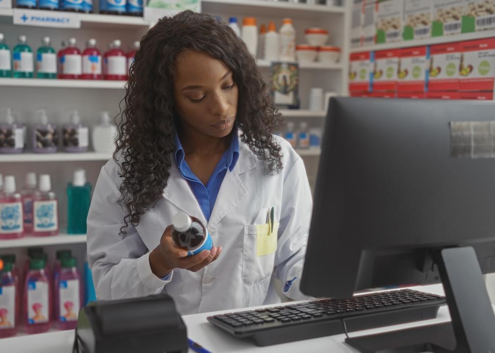 A pharmacist stands in front of a computer in a pharmacy, examining a bottle of medicine.