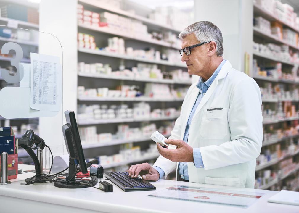 In a pharmacy, a pharmacist stands in front of a computer, holding a box of medication.