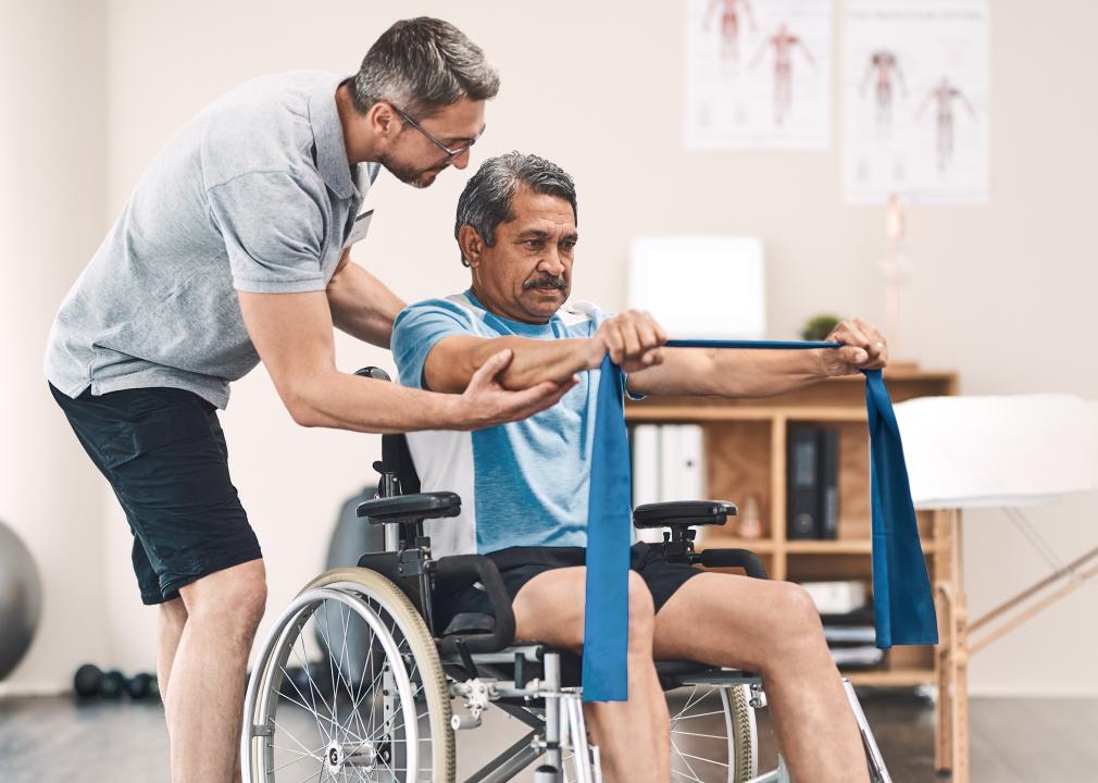 A physiotherapist helps a patient in a wheelchair holding a stretchy band out in front of them.