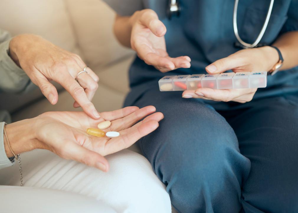 A person in scrubs holding a pill box sitting next to a person on a sofa holding a few pills in her hand.