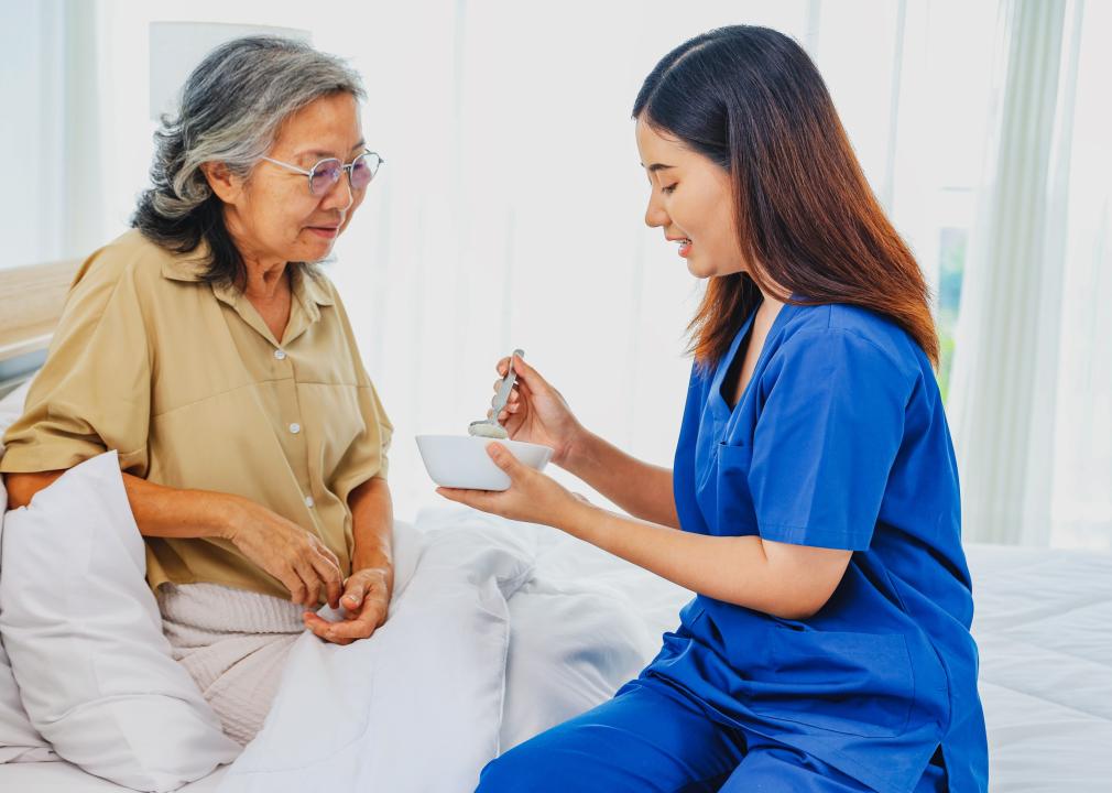 A nursing assistant in scrubs feeds an older woman seated in a bed.