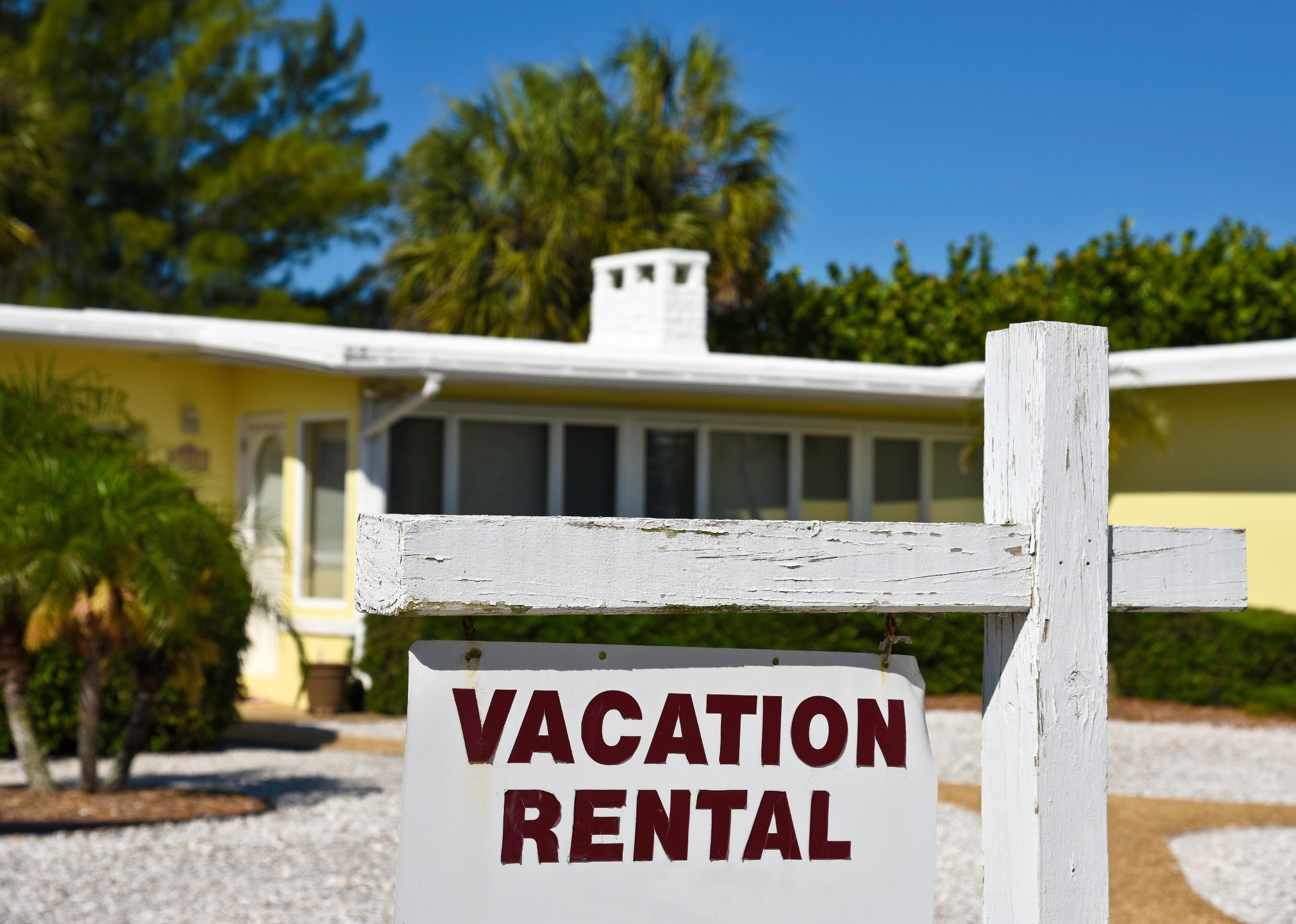 A vacation rental sign in front of a yellow one story home on the beach.