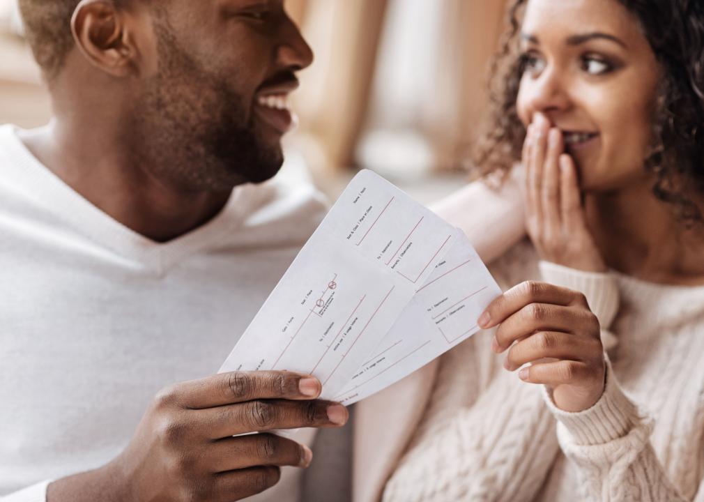 A man hands airline tickets to a woman who is smiling with her hand over her mouth. 