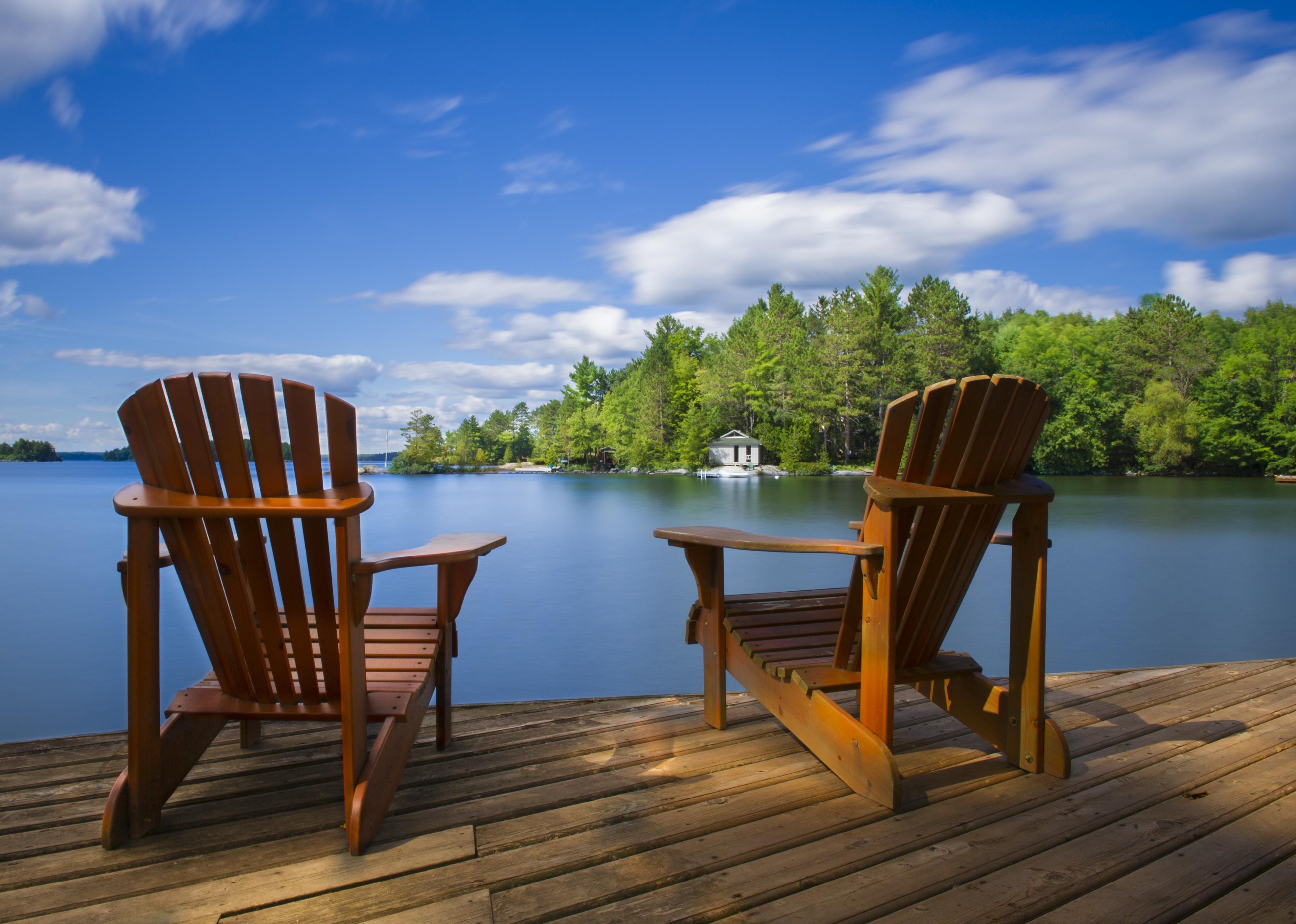 Two Muskoka chairs sitting on a wood dock facing a calm lake.