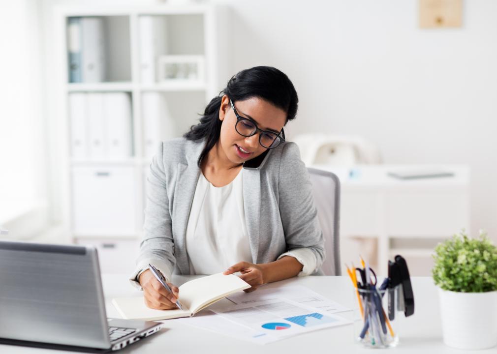 A women sitting behind a computer, writes in a notebook and speaks on the phone. 