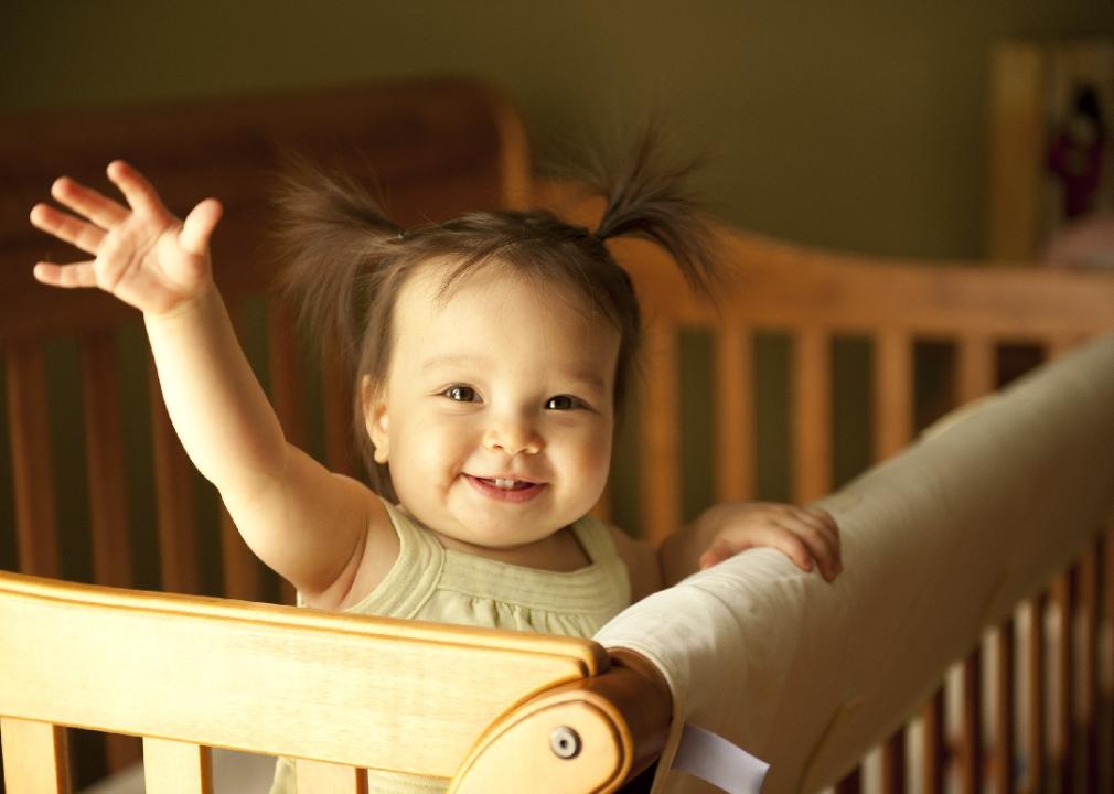 Baby girl waving hand and standing up in crib.