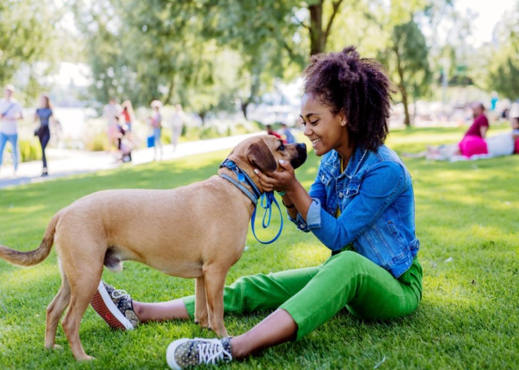 A smiling girl sitting on the grass, lovingly holding a medium-sized dog's head in her hands. 