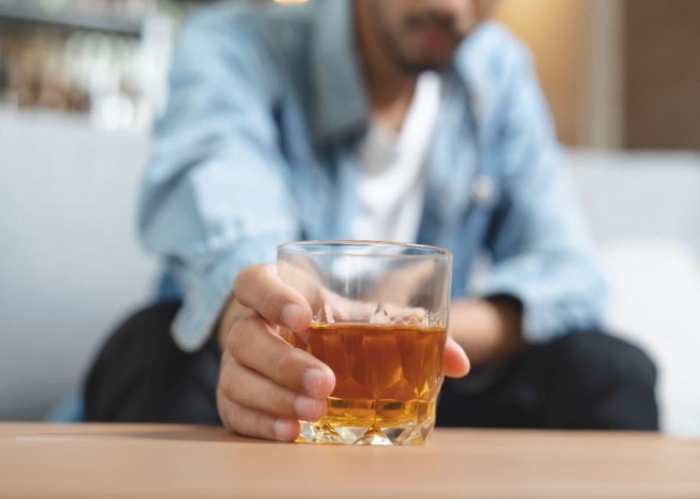 A man reaching for a glass of brown liquor on a table.