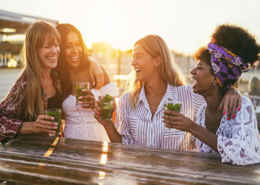 A group of women laughing and drinking cocktails outside as the sun is setting. 