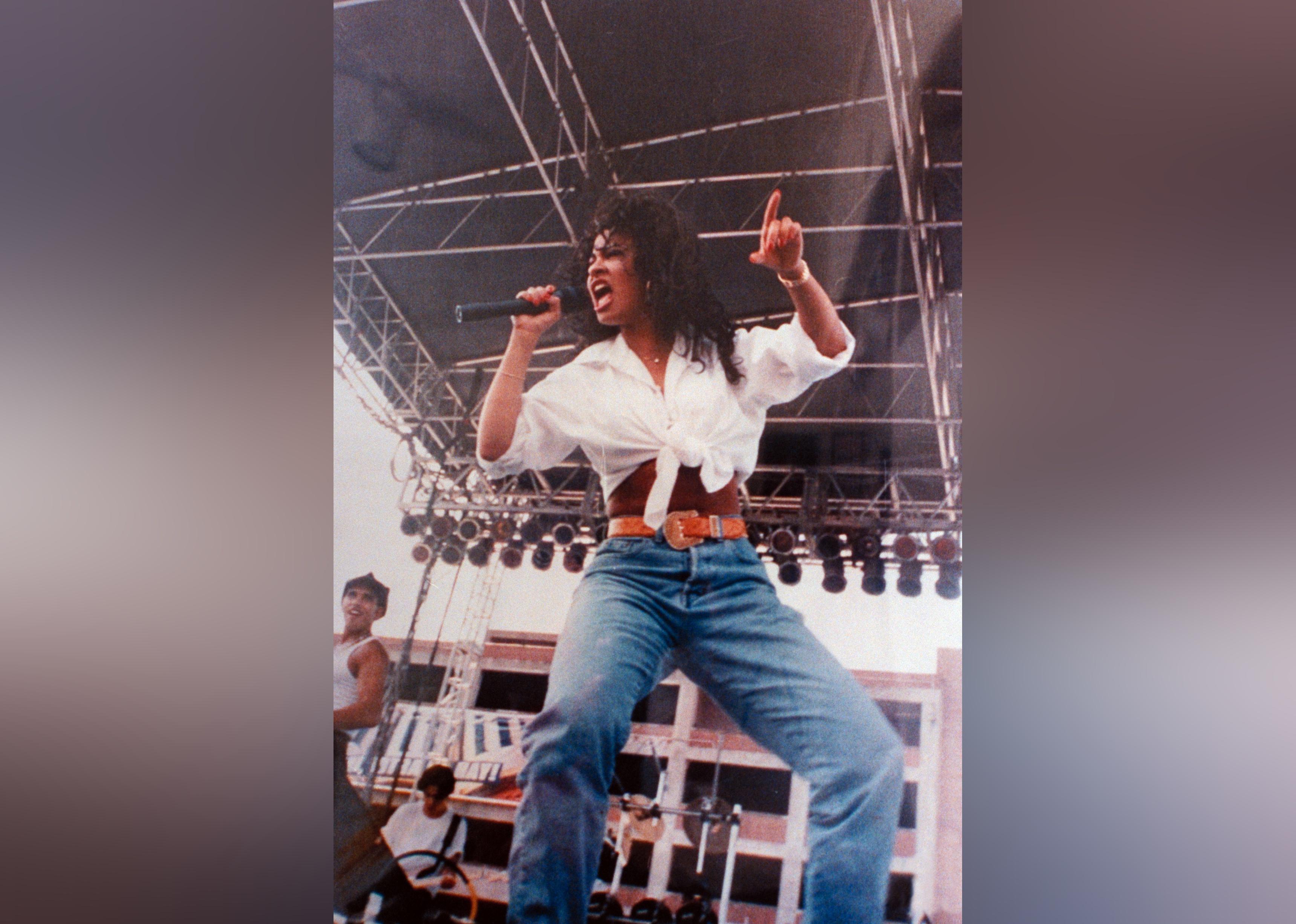 Tejano singer Selena singing into mike on outdoor stage. 