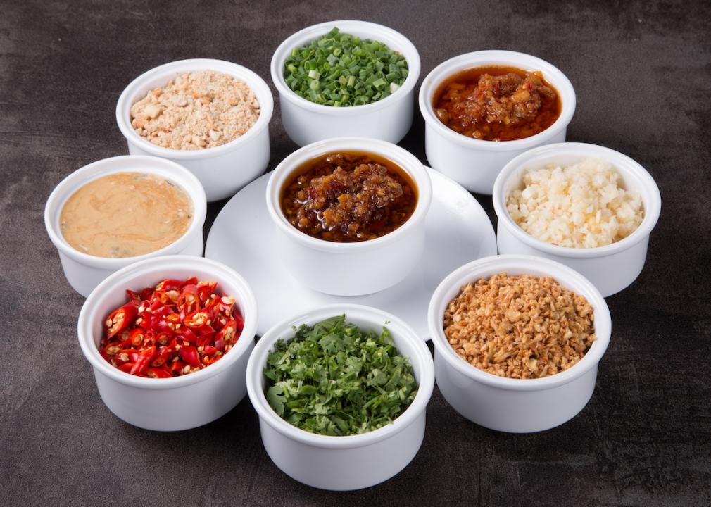 Nine ramekins of ingredients that go into hot, including crushed peanuts, fresh chives, fresh cilantro, fresh garlic, fresh chillies, and various pastes and sauces.arranged in a circle on a table.