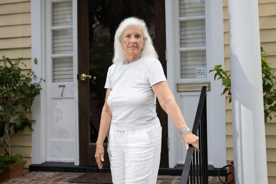 Susan Lyons, an older woman with long white hair, stands in front of her beige and white house holding onto the black handrail.